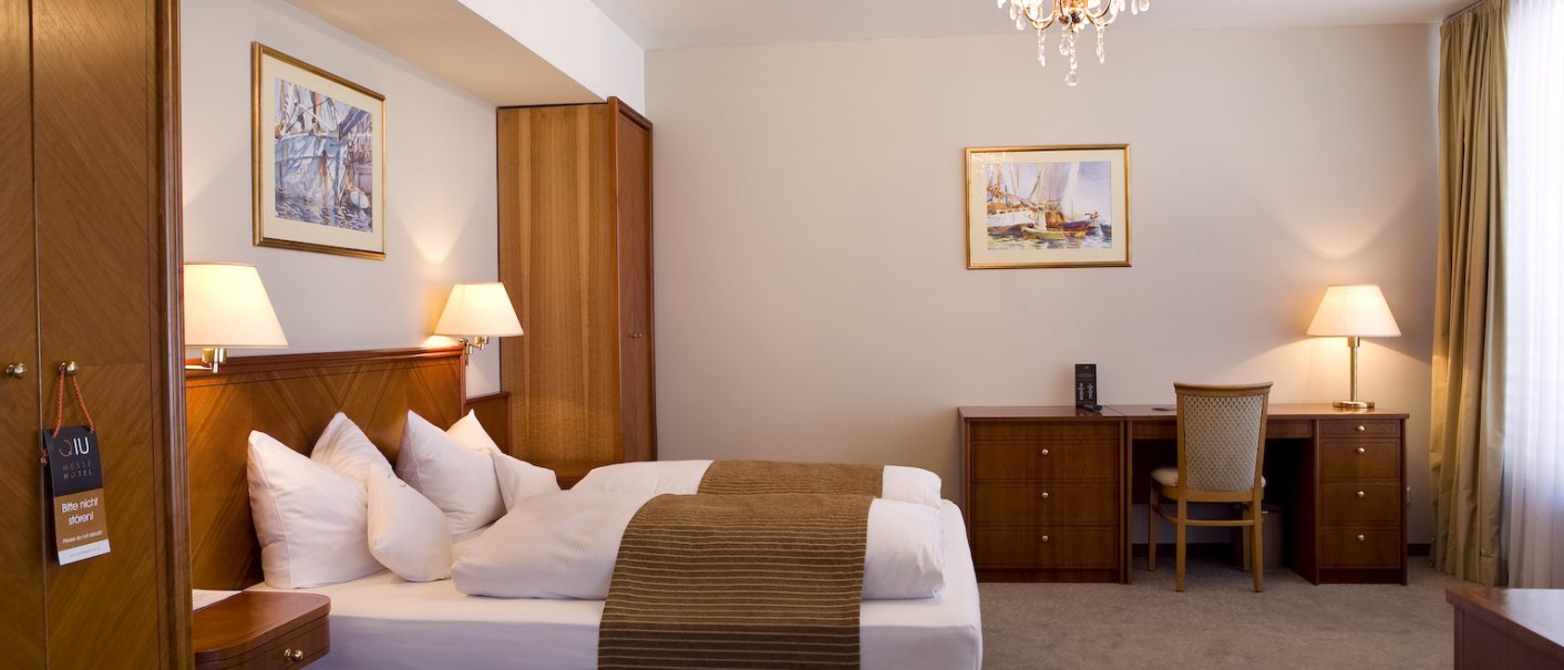 Elegantes Doppelzimmer mit Doppelbett, Schreibtisch, Kronleuchter und zwei Lampen. Warme Holzmöbel und Bilder an den Wänden., © Ross Messehotel Elegantes Doppelzimmer mit Doppelbett, Schreibtisch, Kronleuchter und zwei Lampen. Warme Holzmöbel und Bilder an den Wänden., © Ross Messehotel