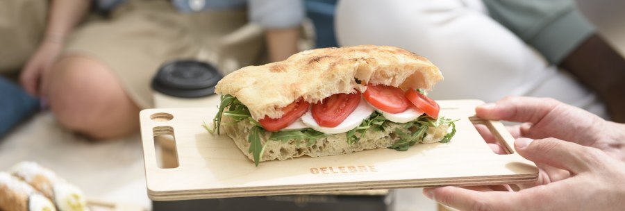 A sandwich with tomatoes, mozzarella and rocket on a wooden board, held by a hand. People having a picnic can be seen in the background., © CELEBRE A sandwich with tomatoes, mozzarella and rocket on a wooden board, held by a hand. People having a picnic can be seen in the background., © CELEBRE