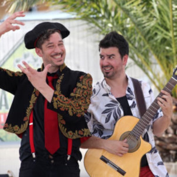 Zwei Männer, einer mit Gitarre, der andere in spanischer Kleidung, gestikulieren fröhlich vor Palmen., © Theaterhaus Stuttgart e.V.