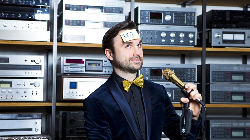 Ein Mann im Anzug mit goldener Fliege hält ein Mikrofon. Auf seiner Stirn klebt ein Zettel mit 'POP'. Im Hintergrund sind viele Audiogeräte zu sehen., © Theaterhaus Stuttgart e.V. Ein Mann im Anzug mit goldener Fliege hält ein Mikrofon. Auf seiner Stirn klebt ein Zettel mit 'POP'. Im Hintergrund sind viele Audiogeräte zu sehen., © Theaterhaus Stuttgart e.V.