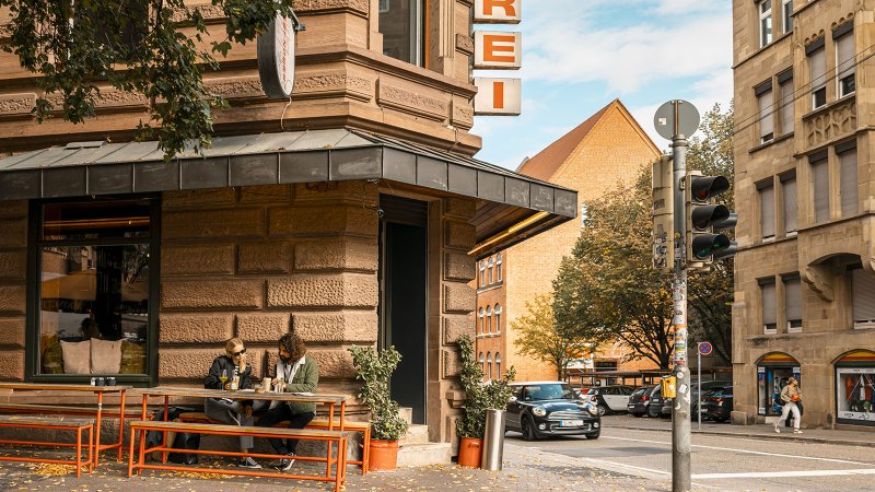 Eine Stra&szlig;enecke mit einer Metzgerei. Zwei Personen sitzen an einem Tisch vor dem Laden. Ein Auto f&auml;hrt vorbei, und es gibt eine Ampel., &copy; SMG, Sarah Schmid