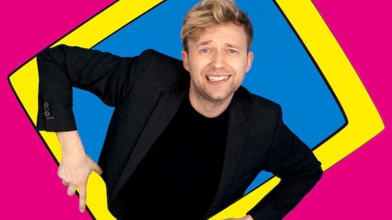 A man in a black suit smiles through a yellow frame on a blue background, surrounded by a pink background., © Renitenztheater Stuttgart e.V. A man in a black suit smiles through a yellow frame on a blue background, surrounded by a pink background., © Renitenztheater Stuttgart e.V.
