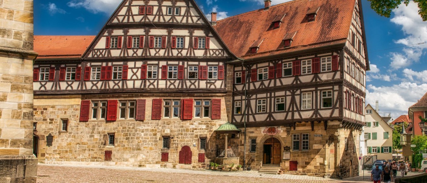 Historisches Fachwerkhaus mit roten Fensterläden und steinernen Details in Esslingen, umgeben von Kopfsteinpflaster und Passanten., © KESSLER Sekt GmbH & Co. KG Historisches Fachwerkhaus mit roten Fensterläden und steinernen Details in Esslingen, umgeben von Kopfsteinpflaster und Passanten., © KESSLER Sekt GmbH & Co. KG