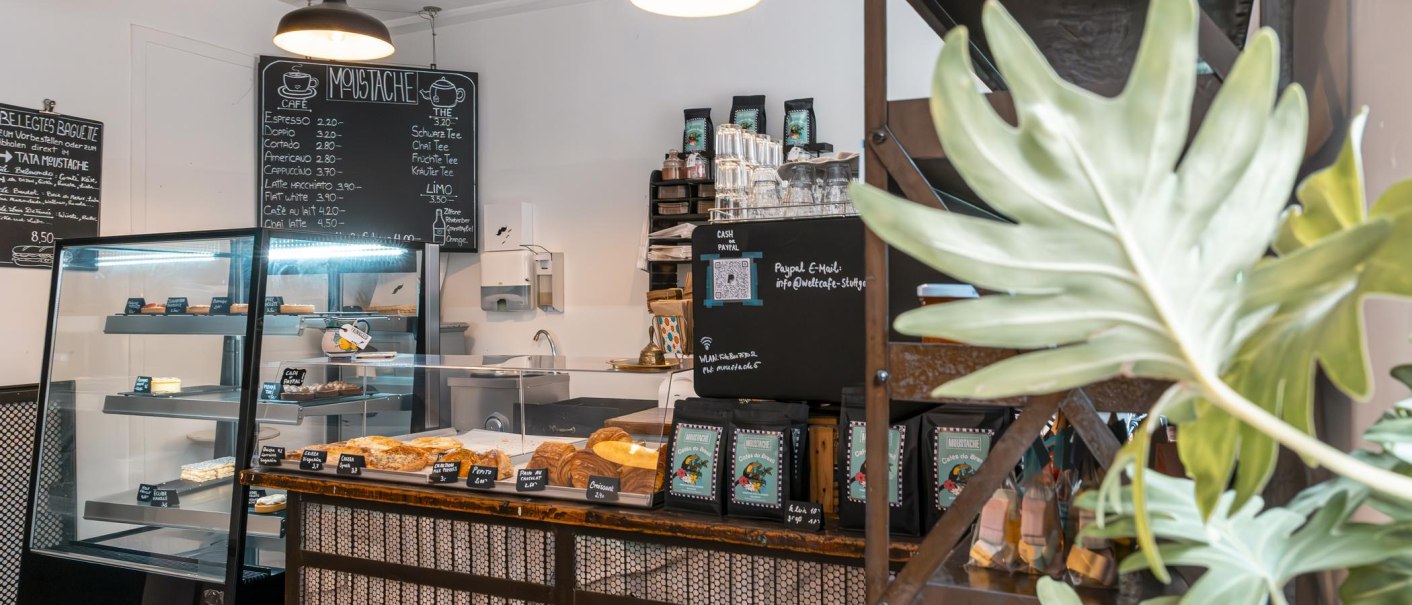 Ein gemütliches Café mit Theke, Gebäckauslage und Getränkekarte. Pflanzen schmücken den Raum, und eine Tafel zeigt das Menü an., © SMG, Sarah Schmid