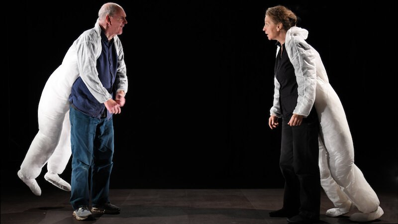 Zwei Personen in weißen, skurrilen Kostümen stehen sich auf einer dunklen Bühne gegenüber und interagieren miteinander., © FITZ Das Theater animierter Formen Zwei Personen in weißen, skurrilen Kostümen stehen sich auf einer dunklen Bühne gegenüber und interagieren miteinander., © FITZ Das Theater animierter Formen