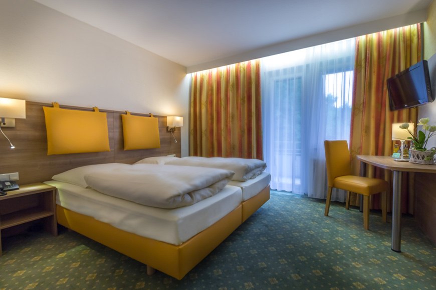 A cozy double room with two beds, a table, a chair, and a TV. Light-colored curtains and warm colors create a welcoming atmosphere., &copy; Kulinarium an der Glems