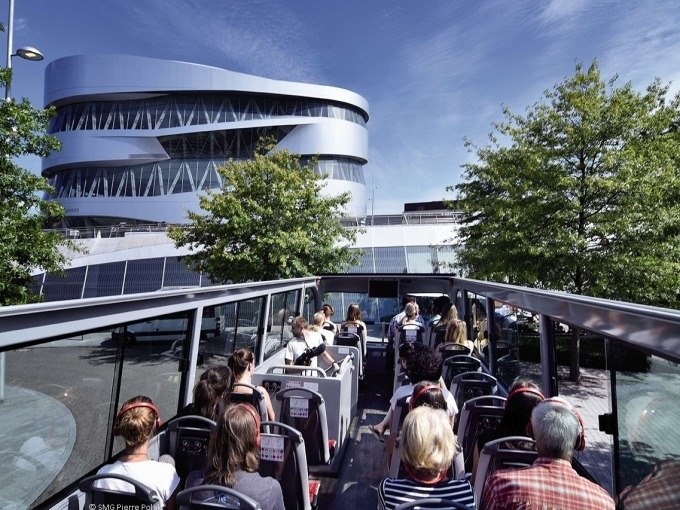 Touristen sitzen in einem offenen Doppeldeckerbus vor dem modernen Mercedes-Benz Museum in Stuttgart, umgeben von B&auml;umen und blauem Himmel., &copy; SMG, Pierre Polak