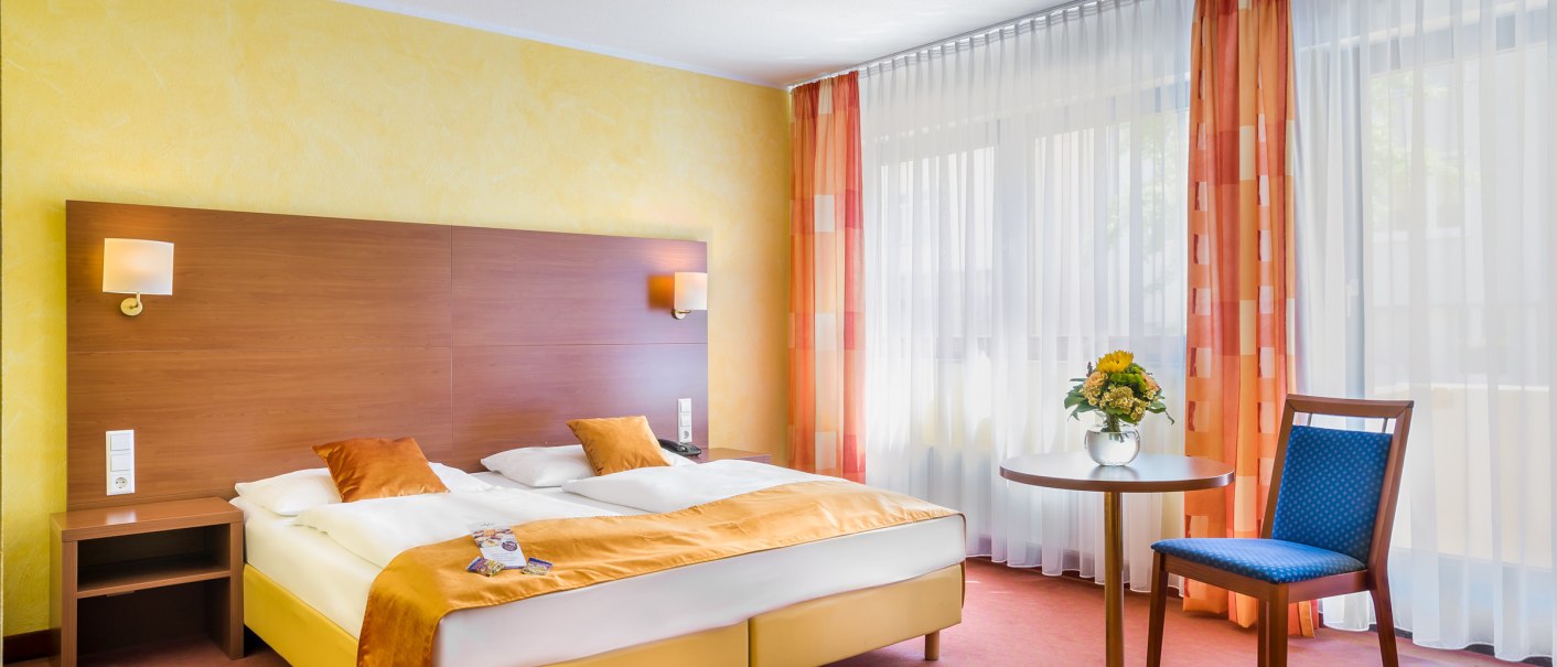 A cozy hotel room with a double bed, yellow walls and orange curtains. A table with flowers and a blue chair complete the ambience., © TOMAS