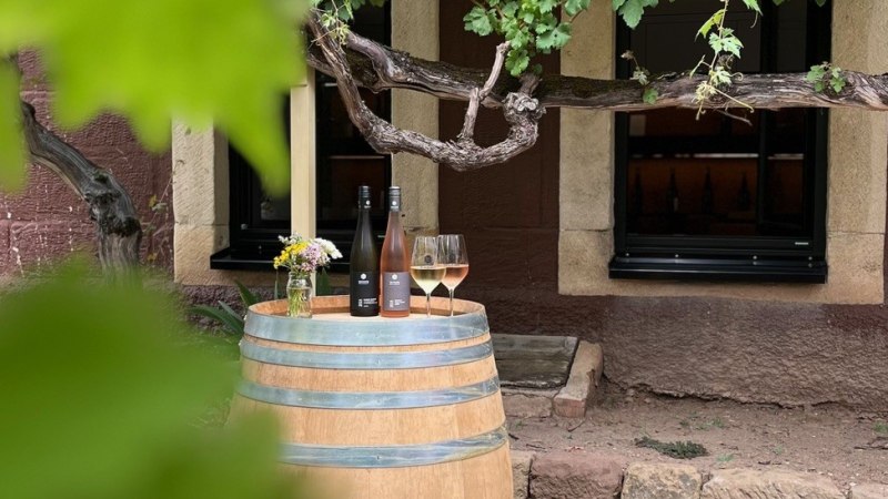 Ein Weinfass dient als Tisch mit zwei Weinflaschen und Gläsern darauf, umgeben von Weinreben vor einem rustikalen Gebäude., © Weingut Schwarz Ein Weinfass dient als Tisch mit zwei Weinflaschen und Gläsern darauf, umgeben von Weinreben vor einem rustikalen Gebäude., © Weingut Schwarz