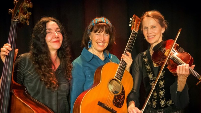 Drei Musikerinnen stehen lächelnd auf einer Bühne. Sie halten einen Kontrabass, eine Gitarre und eine Geige und strahlen Freude aus., © Maslband Drei Musikerinnen stehen lächelnd auf einer Bühne. Sie halten einen Kontrabass, eine Gitarre und eine Geige und strahlen Freude aus., © Maslband