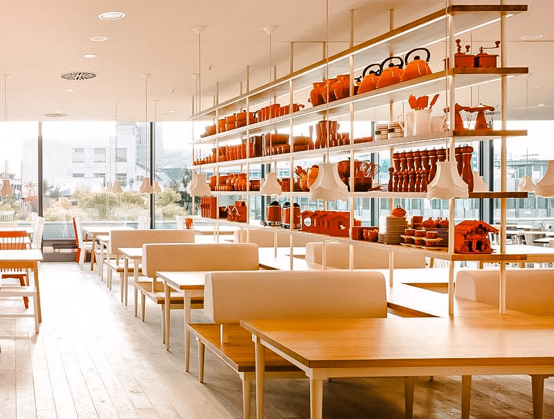 Modernes Restaurant mit weißen Tischen und Regalen voller roter Dekorationen. Große Fenster bieten Blick nach draußen., © Breuninger