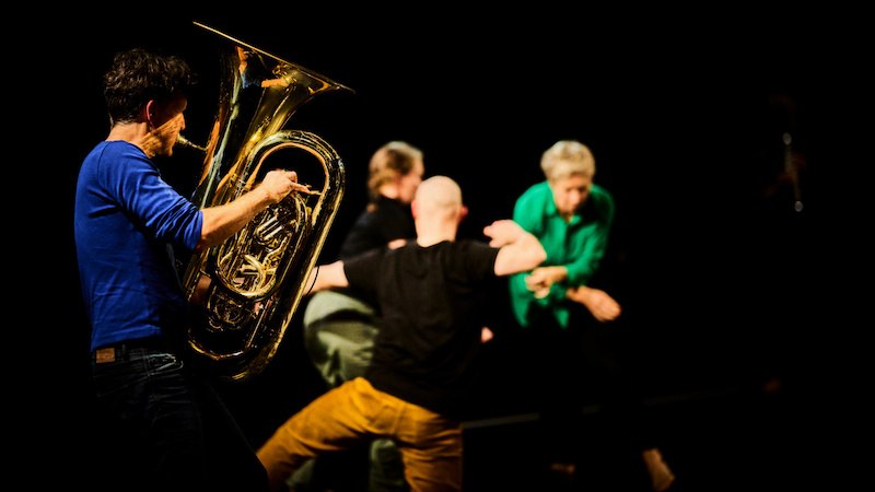 Ein Tubaspieler in blauem Shirt spielt leidenschaftlich, während im Hintergrund Tänzer in dynamischen Posen zu sehen sind., © FITZ Das Theater animierter Formen Ein Tubaspieler in blauem Shirt spielt leidenschaftlich, während im Hintergrund Tänzer in dynamischen Posen zu sehen sind., © FITZ Das Theater animierter Formen