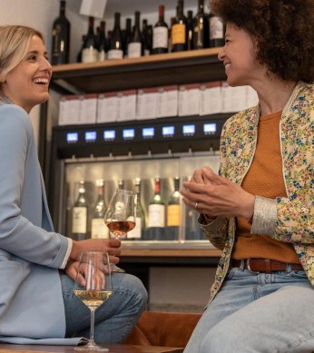 Zwei Frauen sitzen auf hohen Stühlen und lachen einander zu. Eine hält in der Hand ein Weinglas. Im Hintergrund ein Weinspender mit zapfbaren Weinflaschen., © Stuttgart-Marketing GmbH, Martina Denker