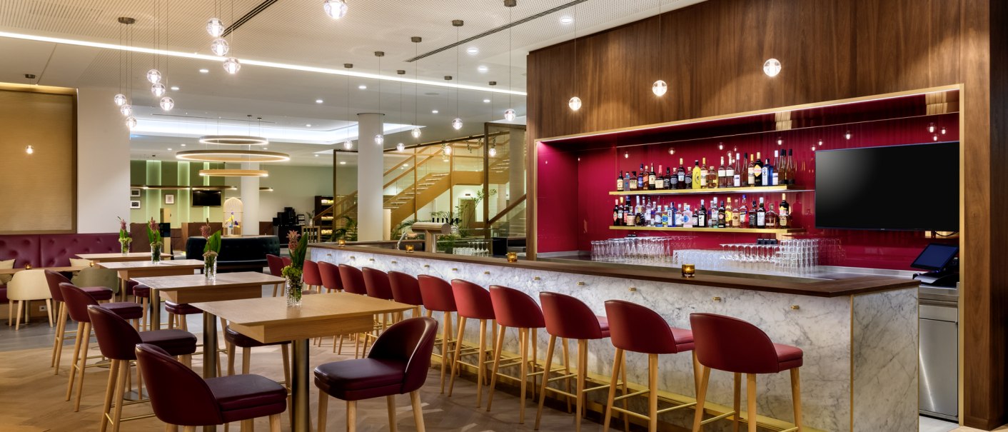 Moderne Bar mit roten Stühlen, Marmor-Theke und beleuchtetem Regal mit Spirituosen. Helle Deckenbeleuchtung und Holzboden schaffen eine einladende Atmosphäre., © Mövenpick Hotel Stuttgart Messe & Congress