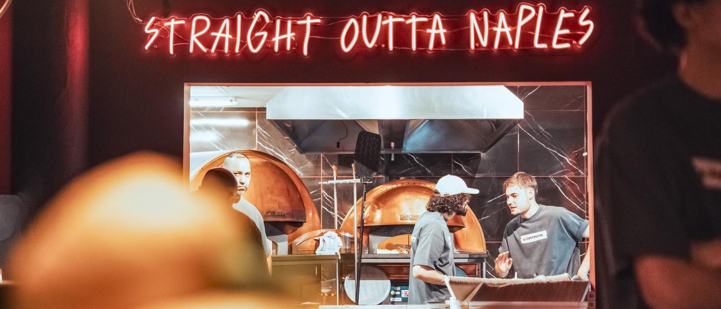 Neon-Schriftzug „STRAIGHT OUTTA NAPLES“ über einer offenen Küche; drei Personen arbeiten in der Küche, vermutlich bei einer L’Artista-Eröffnung., © L'Artista Stuttgart GmbH Neon-Schriftzug „STRAIGHT OUTTA NAPLES“ über einer offenen Küche; drei Personen arbeiten in der Küche, vermutlich bei einer L’Artista-Eröffnung., © L'Artista Stuttgart GmbH