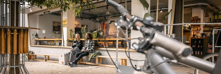Zwei Personen sitzen lachend vor einem Caf&eacute; auf einer Bank. Im Vordergrund ist ein Fahrradlenker zu sehen. Herbstliche Bl&auml;tter liegen auf dem Boden., &copy; SMG Stuttgart Marketing GmbH - Sarah Schmid