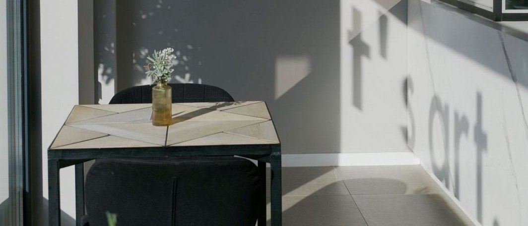 Heller Raum mit Tisch, zwei schwarzen Stühlen und einer kleinen Vase; an der Wand fällt der Schatten des Wortes „Short“., © Taraba Heller Raum mit Tisch, zwei schwarzen Stühlen und einer kleinen Vase; an der Wand fällt der Schatten des Wortes „Short“., © Taraba