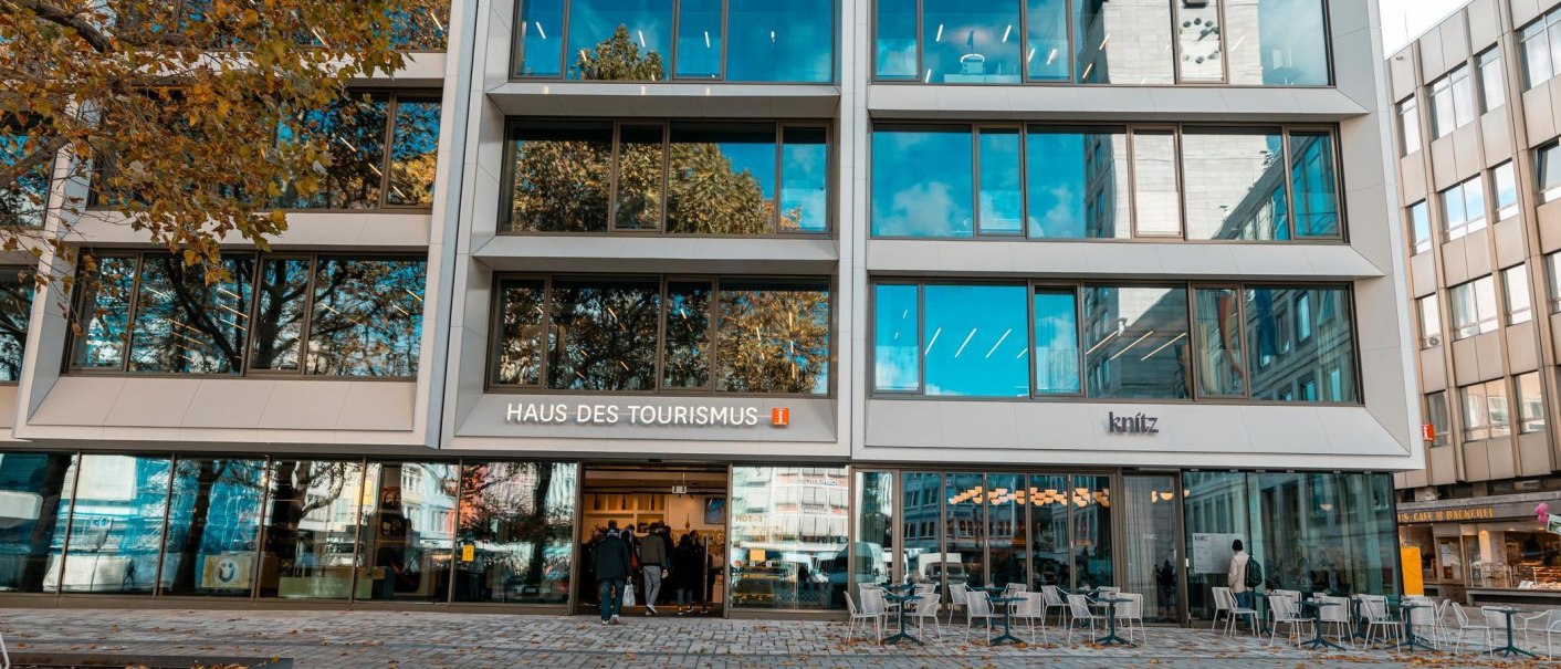 Moderne Glasfassade des Haus des Tourismus mit Spiegelungen von Bäumen und blauem Himmel. Menschen betreten die Tourist-Information im Gebäude, draußen stehen Tische und Stühle., © SMG, Sarah Schmid Moderne Glasfassade des Haus des Tourismus mit Spiegelungen von Bäumen und blauem Himmel. Menschen betreten die Tourist-Information im Gebäude, draußen stehen Tische und Stühle., © SMG, Sarah Schmid