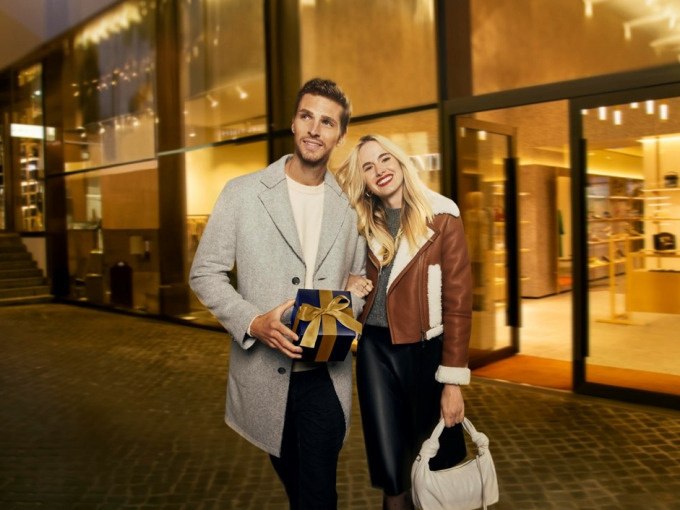 Ein Paar spaziert abends vor einem beleuchteten Geschäft. Der Mann hält ein Geschenk, die Frau lächelt und trägt eine Handtasche., © Schlienz-Tours GmbH & Co.KG Ein Paar spaziert abends vor einem beleuchteten Geschäft. Der Mann hält ein Geschenk, die Frau lächelt und trägt eine Handtasche., © Schlienz-Tours GmbH & Co.KG