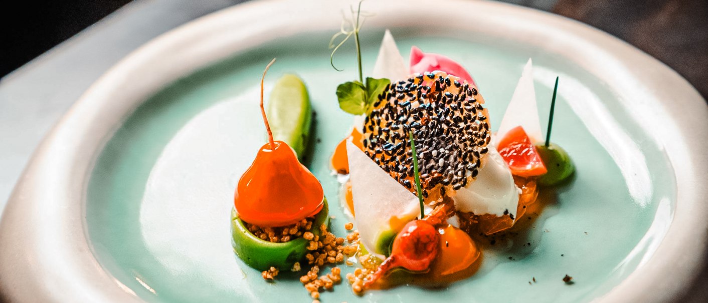 Ein kunstvoll angerichtetes Gourmetgericht auf einem Teller mit grüner Glasur. Verschiedene bunte Zutaten und dekorative Elemente sind zu sehen., © wosilatfotografie Ein kunstvoll angerichtetes Gourmetgericht auf einem Teller mit grüner Glasur. Verschiedene bunte Zutaten und dekorative Elemente sind zu sehen., © wosilatfotografie