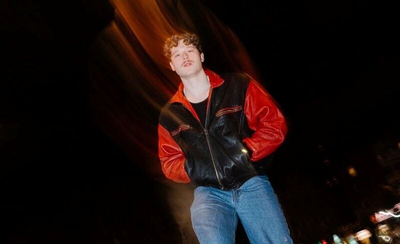 A man in a black and red leather jacket stands on a street at night. The background is blurred, suggesting dynamic movement., © Marvin Hoffmann A man in a black and red leather jacket stands on a street at night. The background is blurred, suggesting dynamic movement., © Marvin Hoffmann