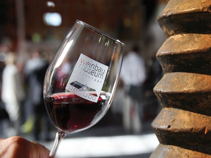 A wine glass with the logo of the Stuttgart Wine Museum, filled with red wine, in front of a blurred background with people., © Stuttgart Marketing GmbH A wine glass with the logo of the Stuttgart Wine Museum, filled with red wine, in front of a blurred background with people., © Stuttgart Marketing GmbH