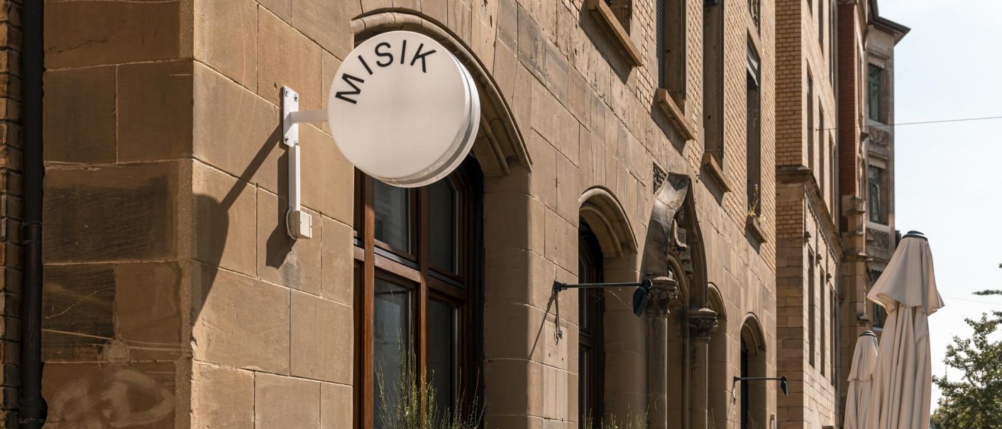 Gebäude mit Sandsteinfassade, rundes Schild mit 'MISIK', Fenster mit Pflanzen, geschlossene Sonnenschirme auf der Straße., © Stuttgart-Marketing GmbH, Sarah Schmid