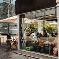 Außenansicht eines Cafés mit Glasfront, Menschen sitzen an Tischen, moderne Architektur im Hintergrund., © SMG, Sarah Schmid