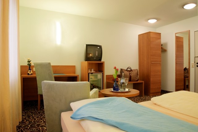 Gemütliches Hotelzimmer mit Doppelbett, Schreibtisch, Fernseher, Minibar und Tisch mit Blumen. Helle Wände und Teppichboden., © ateckhotel
