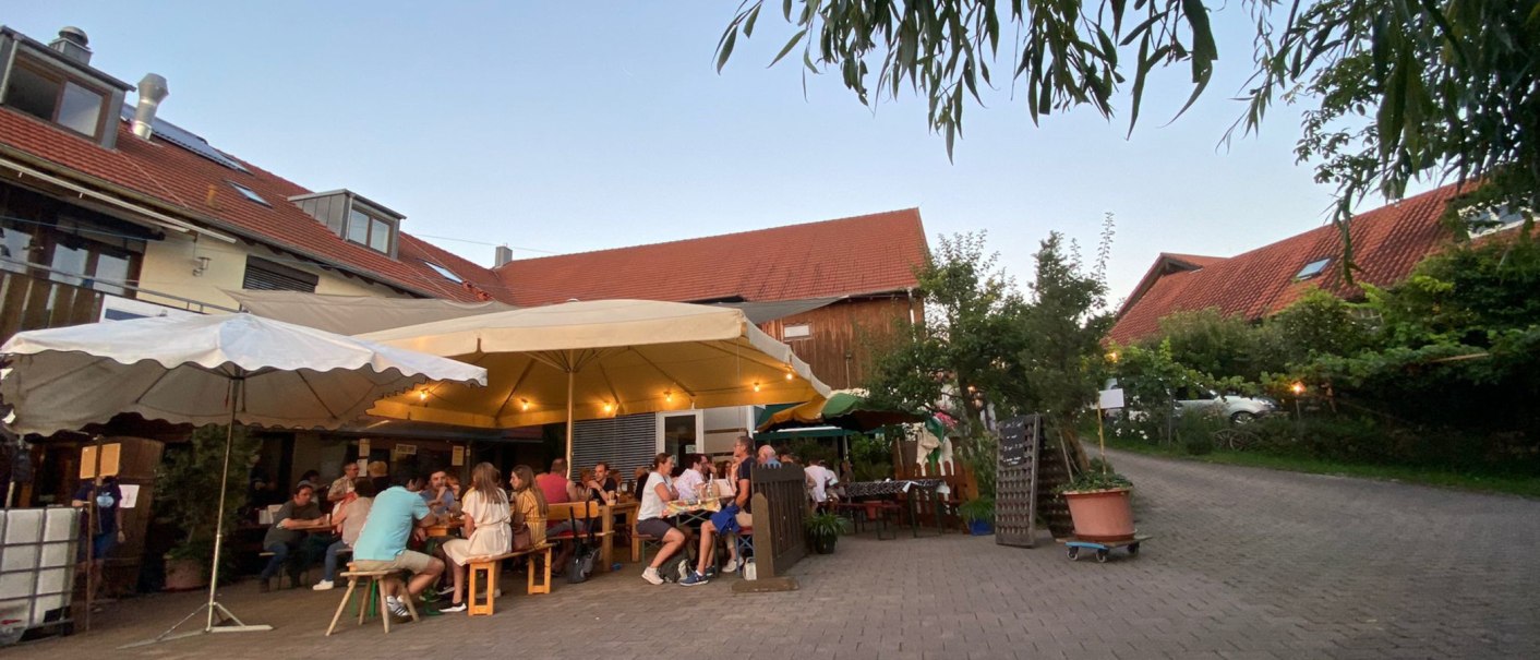 Menschen sitzen unter großen Sonnenschirmen in einem gemütlichen Hof mit roten Dächern und Bäumen., © Weinhof Zaiß Menschen sitzen unter großen Sonnenschirmen in einem gemütlichen Hof mit roten Dächern und Bäumen., © Weinhof Zaiß
