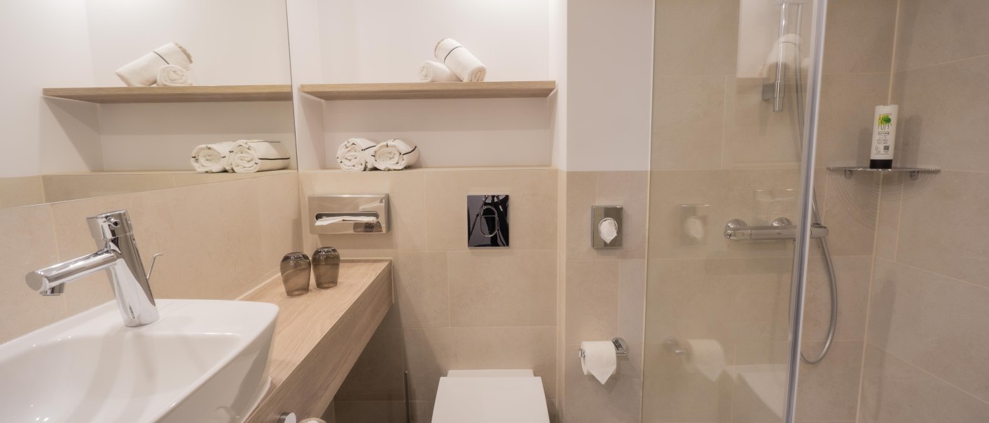 Modernes Badezimmer mit Waschbecken, Toilette, Dusche und Handt&uuml;chern auf Regalen. Helle Fliesen und minimalistische Einrichtung., &copy; HARBR. hotel Ludwigsburg / DQuadrat Living GmbH