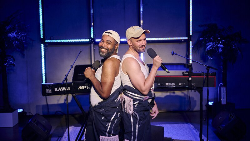 Zwei Männer stehen Rücken an Rücken auf einer Bühne, halten Mikrofone und lächeln. Im Hintergrund sind ein Keyboard und Palmen zu sehen., © Württembergische Staatstheater Stuttgart Zwei Männer stehen Rücken an Rücken auf einer Bühne, halten Mikrofone und lächeln. Im Hintergrund sind ein Keyboard und Palmen zu sehen., © Württembergische Staatstheater Stuttgart