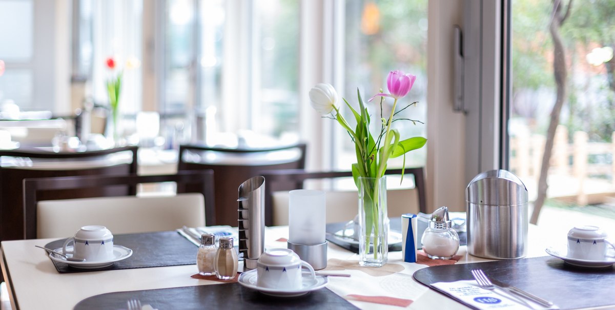 Ein Tisch in einem Caf&eacute; ist mit Geschirr, Besteck und einer Vase mit Tulpen gedeckt. Im Hintergrund sind Fenster und weitere Tische zu sehen., &copy; TOMAS