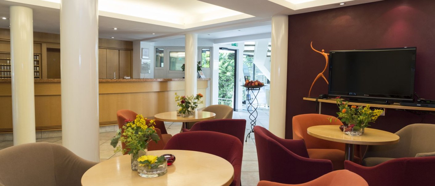Modern hotel lobby with cozy armchairs, tables with flowers, a large TV and a reception desk in the background., © Hotel Gloria GmbH Restaurant Möhringer Hexle Modern hotel lobby with cozy armchairs, tables with flowers, a large TV and a reception desk in the background., © Hotel Gloria GmbH Restaurant Möhringer Hexle