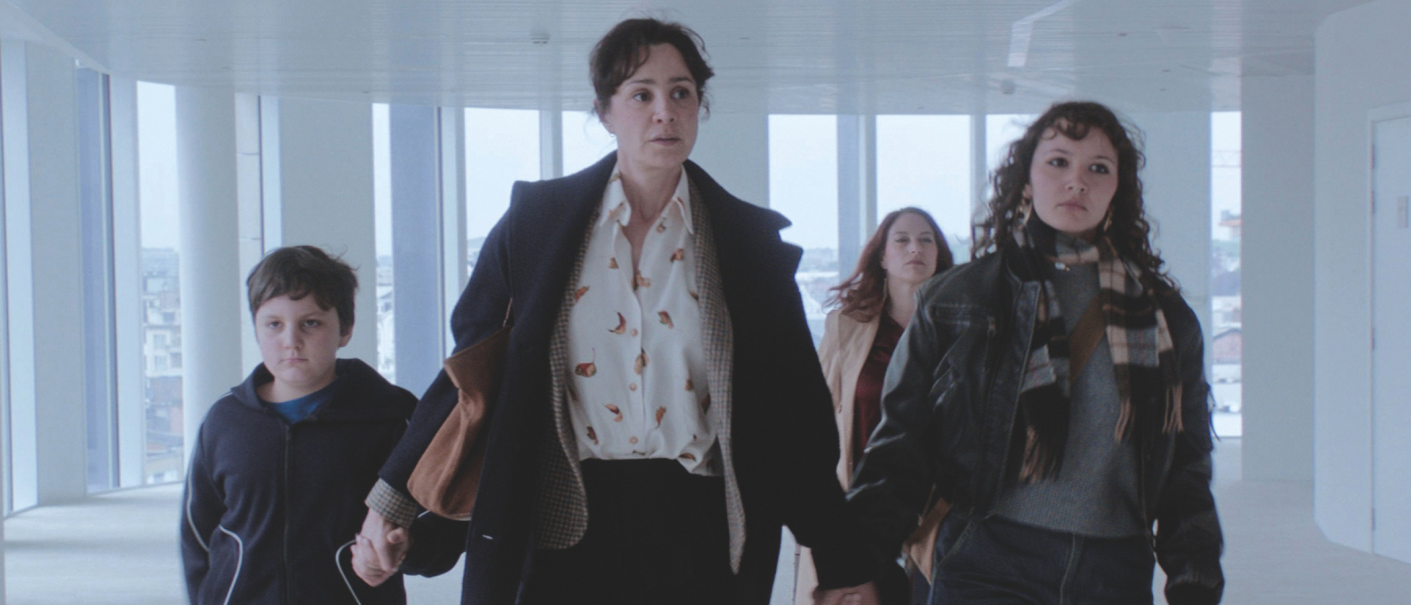 Vier Personen, darunter ein Kind, gehen durch einen modernen, hellen Korridor mit großen Fenstern., © eksystent Filmverleih