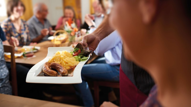 Ein Teller mit geschmorten Bratwürsten, Kartoffelpüree und Salat wird in einem gemütlichen Restaurant serviert. Im Hintergrund sitzen Menschen an einem Holztisch und genießen ihr Essen in geselliger Runde., © SMG, Jean-Claude Winkler Ein Teller mit geschmorten Bratwürsten, Kartoffelpüree und Salat wird in einem gemütlichen Restaurant serviert. Im Hintergrund sitzen Menschen an einem Holztisch und genießen ihr Essen in geselliger Runde., © SMG, Jean-Claude Winkler
