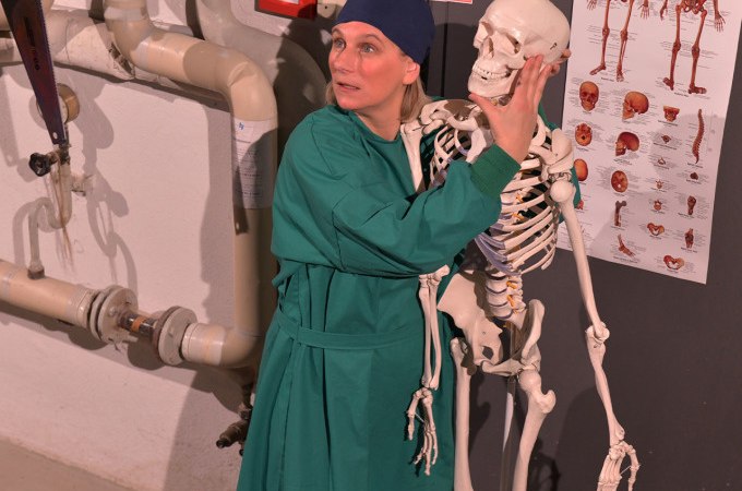 Person in green clothing hugs a skeleton in a room with medical posters and pipes on the wall., © Landesmuseum Württemberg Person in green clothing hugs a skeleton in a room with medical posters and pipes on the wall., © Landesmuseum Württemberg