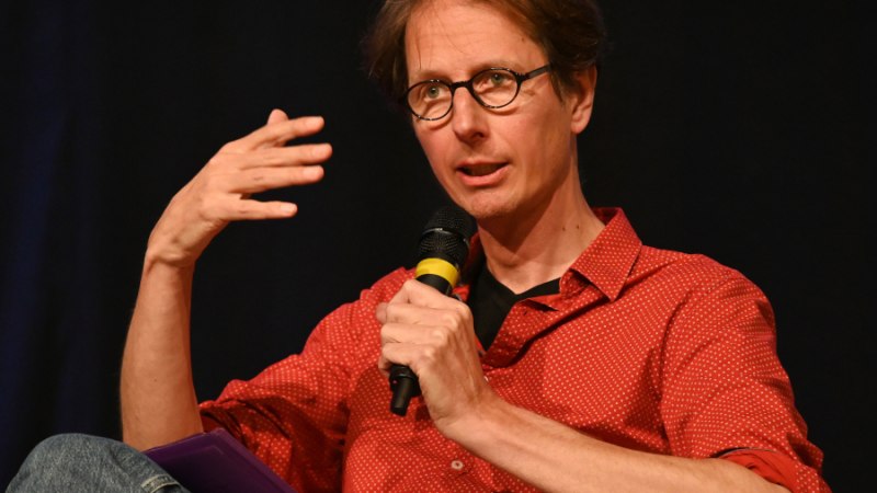 Eine Person in einem roten Hemd spricht auf einer Bühne in ein Mikrofon. Sie gestikuliert mit der linken Hand und trägt eine Brille., © Foto: Günter Giese Eine Person in einem roten Hemd spricht auf einer Bühne in ein Mikrofon. Sie gestikuliert mit der linken Hand und trägt eine Brille., © Foto: Günter Giese