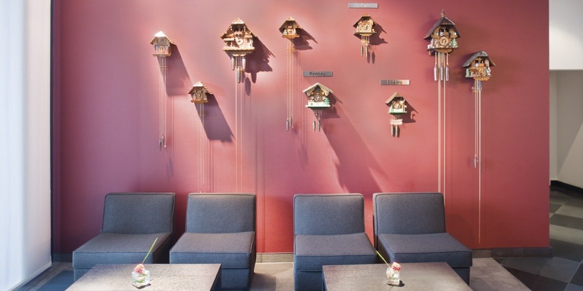 Rote Wand mit mehreren Kuckucksuhren, darunter graue Sitzmöbel. Die Uhren sind mit Städtenamen beschriftet., © Mövenpick Hotel Stuttgart Airport Rote Wand mit mehreren Kuckucksuhren, darunter graue Sitzmöbel. Die Uhren sind mit Städtenamen beschriftet., © Mövenpick Hotel Stuttgart Airport