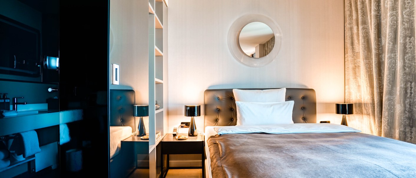 Modernes Hotelzimmer mit elegantem Doppelbett, runden Spiegel über dem Kopfteil, Nachttischen mit Lampen und Vorhängen an der Seite., © CLOUD N°7 APARTMENTS GmbH Modernes Hotelzimmer mit elegantem Doppelbett, runden Spiegel über dem Kopfteil, Nachttischen mit Lampen und Vorhängen an der Seite., © CLOUD N°7 APARTMENTS GmbH