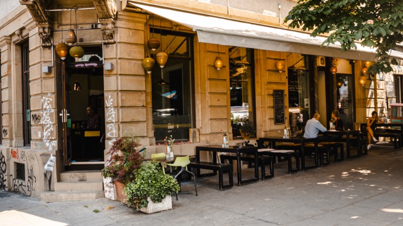 Ein Café an einer Straßenecke mit Außenbestuhlung, Pflanzen und hängenden Lampen. Menschen sitzen an Tischen unter einem Sonnenschutz., © SMG, Sarah Schmid