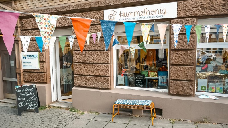 Ladenfront von Hummelhonig mit bunten Wimpeln und einer Bank. Schaufenster zeigt Stoffe und Schnittmuster. Schild der Steuerkanzlei Wanner daneben., © Lena Beck, Hummelkonig GbR, Stuttgart Ladenfront von Hummelhonig mit bunten Wimpeln und einer Bank. Schaufenster zeigt Stoffe und Schnittmuster. Schild der Steuerkanzlei Wanner daneben., © Lena Beck, Hummelkonig GbR, Stuttgart