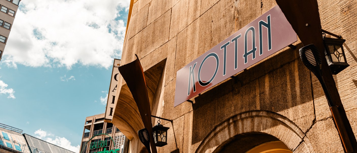 Ein Gebäude mit einem Schild "KOTTAN" und einer Laterne, daneben moderne Gebäude und ein blauer Himmel mit Wolken., © SMG_Sarah Schmid