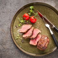 A plate with sliced steak, two cherry tomatoes and cutlery on a rustic base., © MAREDO A plate with sliced steak, two cherry tomatoes and cutlery on a rustic base., © MAREDO