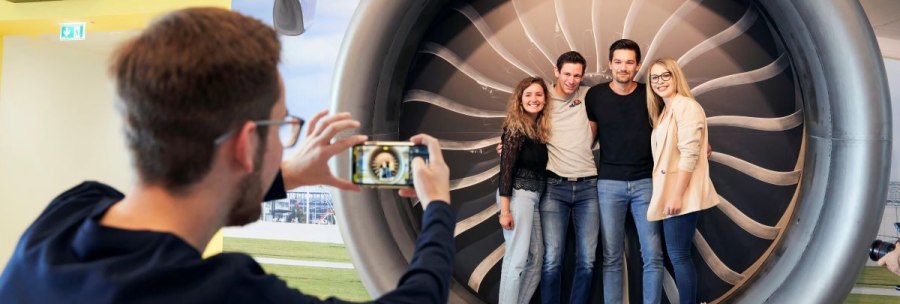 Vier Personen stehen in einer großen Flugzeugturbine, während eine weitere Person ein Foto von ihnen macht. Hintergrund zeigt Flughafenmotiv., © Flughafen Stuttgart