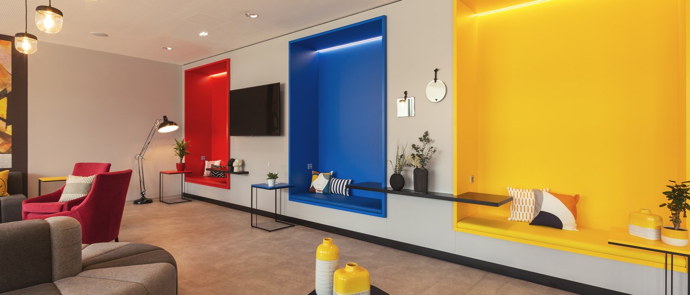 A modern lobby with colorful seating nooks in red, blue, and yellow, decorated with pillows and plants., © the niu Form - Novum Hospitality A modern lobby with colorful seating nooks in red, blue, and yellow, decorated with pillows and plants., © the niu Form - Novum Hospitality