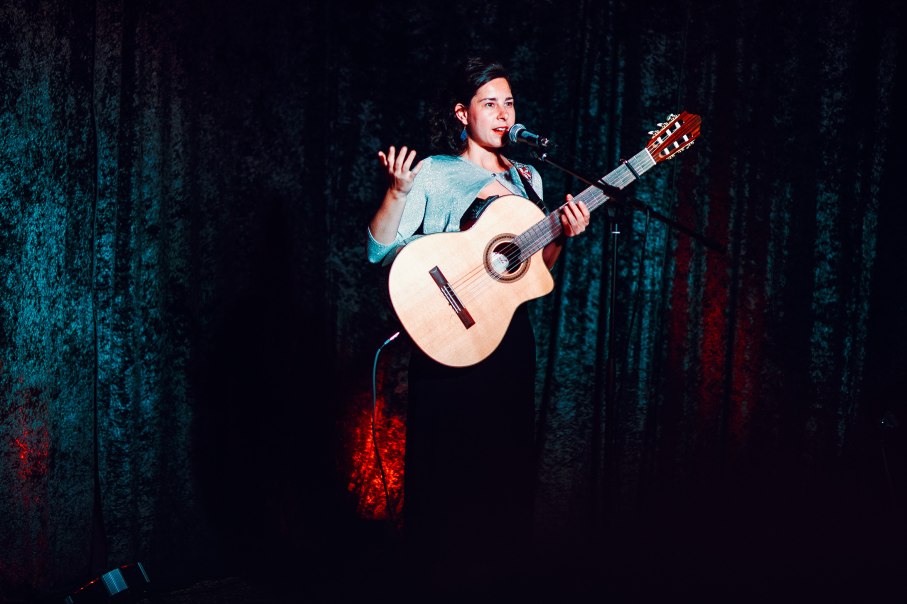 Eine Musikerin steht mit Gitarre auf einer B&uuml;hne und spricht ins Mikrofon. Der Hintergrund ist dunkel mit roter und blauer Beleuchtung., &copy; Paula Linke