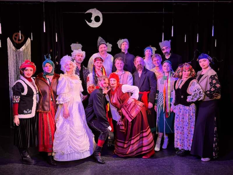 Two people in traditional Ukrainian clothing are standing on a stage, smiling. The man is wearing a cap, and the woman is wearing a red headscarf., © Theater Atelier Two people in traditional Ukrainian clothing are standing on a stage, smiling. The man is wearing a cap, and the woman is wearing a red headscarf., © Theater Atelier