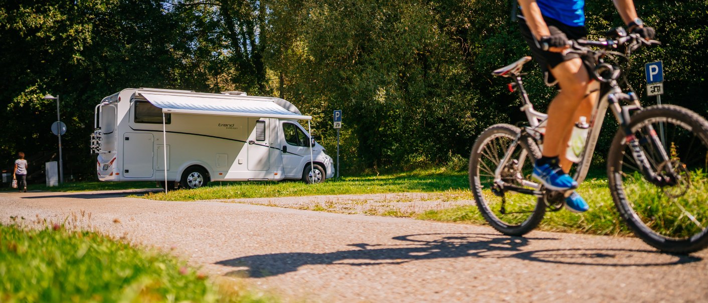 Wohnmobil auf Stellplatz in grüner Umgebung, Radfahrer fährt vorbei. Sonniges Wetter, Bäume im Hintergrund., © SMG, Thomas Niedermüller
