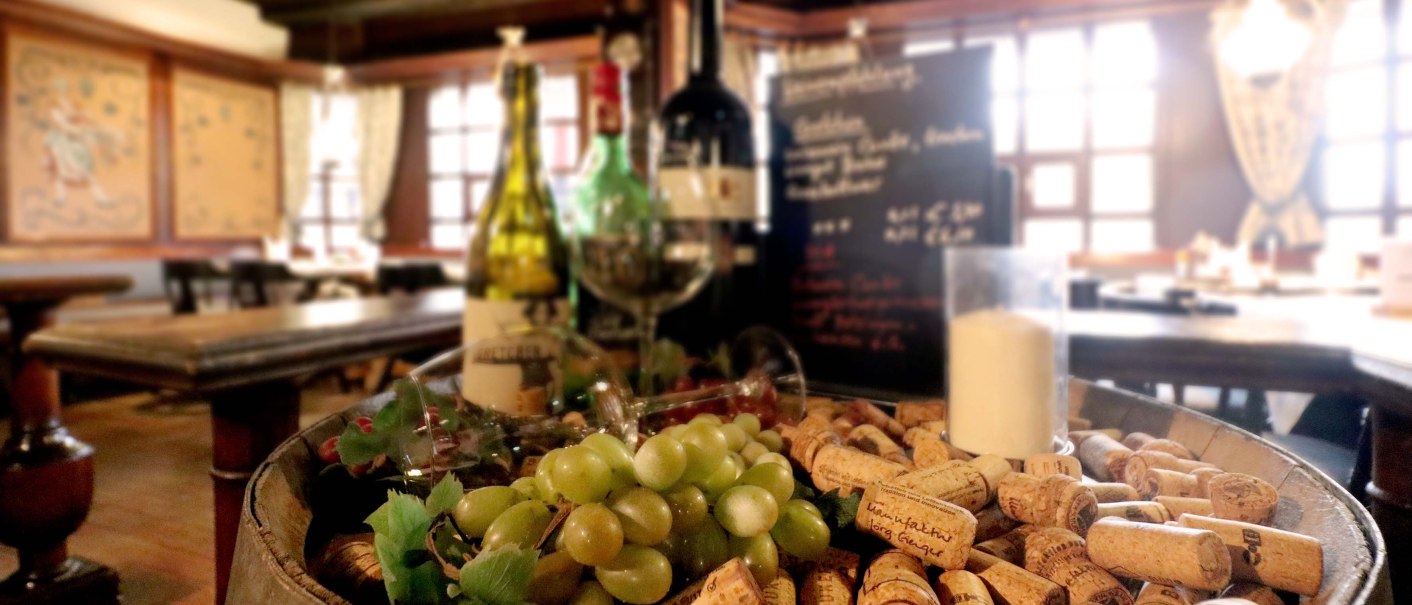 Ein Weinfass mit Korken, Trauben und Weinflaschen in einer gemütlichen Weinstube. Im Hintergrund sind Holztische und eine Tafel zu sehen., © ©hotel-graf-eberhard.de